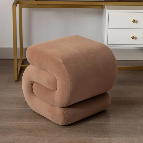 "Angled front view of a brown S-shape ottoman stool with plush cushioning on wooden flooring next to a gold vanity."